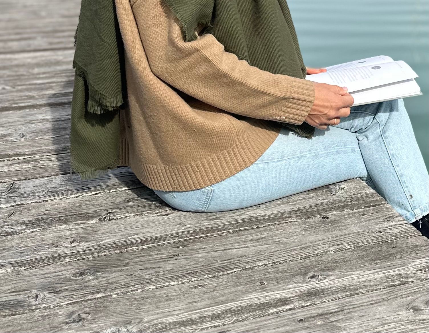 Detail: Hände, aufgeschlagenes Buch, Camel-Pullover, Oliv-Schal, Holz-Steg.